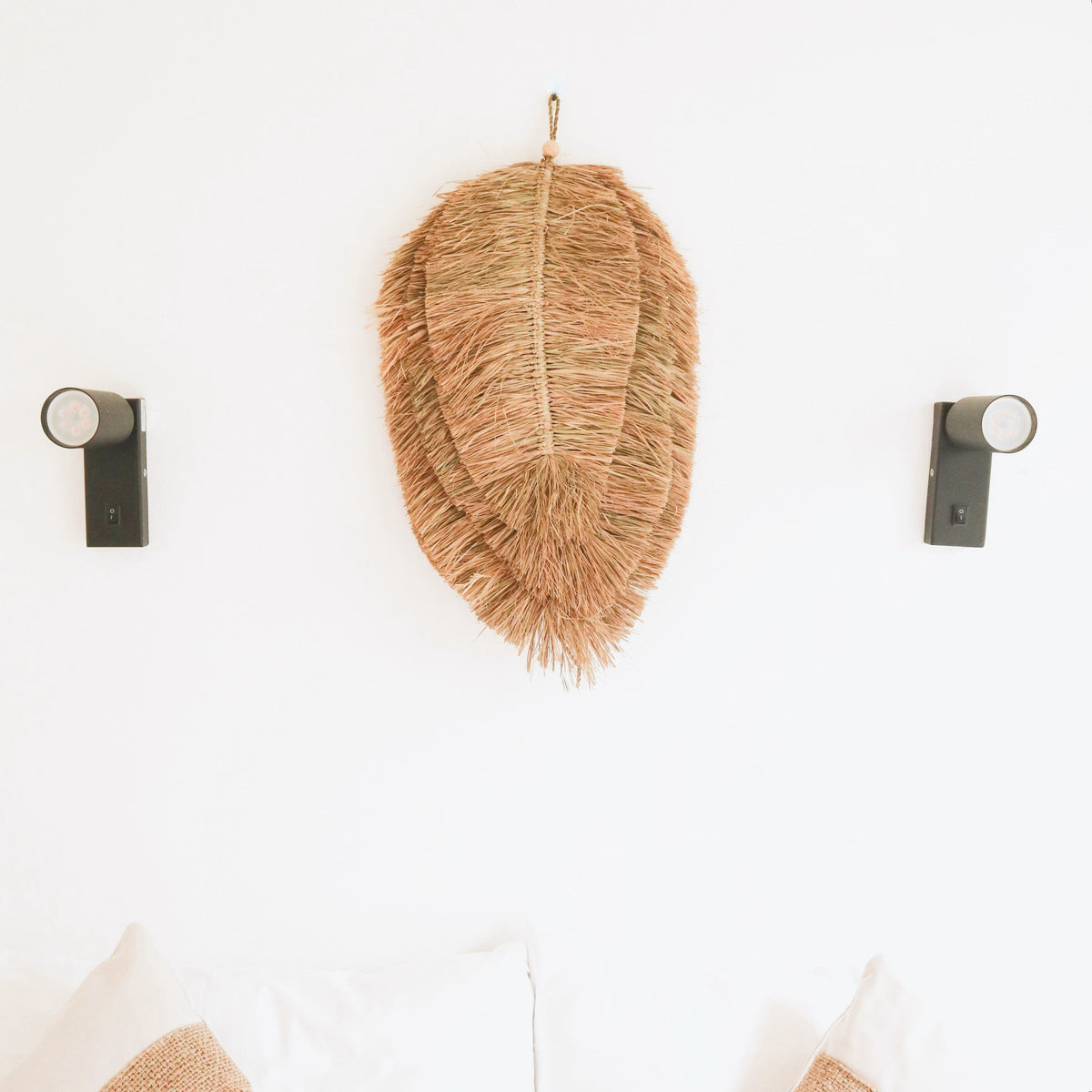 Blattförmige Raffia-Wanddeko TIPIN an weißer Wand über Sofa mit Naturkissen