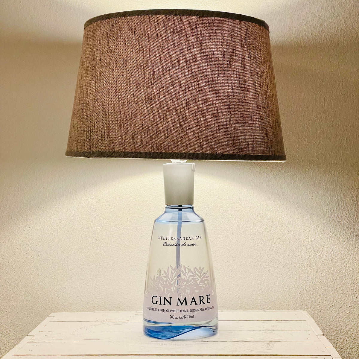 Gin Mare Flaschenlampe aus transparenter Ginflasche mit braunem Stoffschirm, eingeschaltet auf weißem Sideboard
