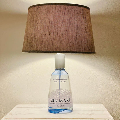 Gin Mare Flaschenlampe aus transparenter Ginflasche mit braunem Stoffschirm, eingeschaltet auf weißem Sideboard