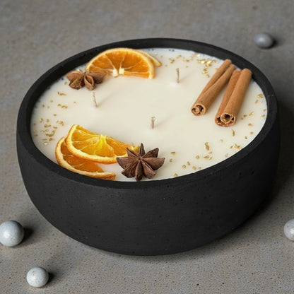 Handgefertigte Duftkerze in schwarzer Beton-Schale mit drei brennenden Dochten, dekoriert mit getrockneten Orangenscheiben, Sternanis, Zimtstangen und goldenen Akzenten – stilvoll inszeniert auf hellem Holztisch mit Vase und Lampe im Hintergrund