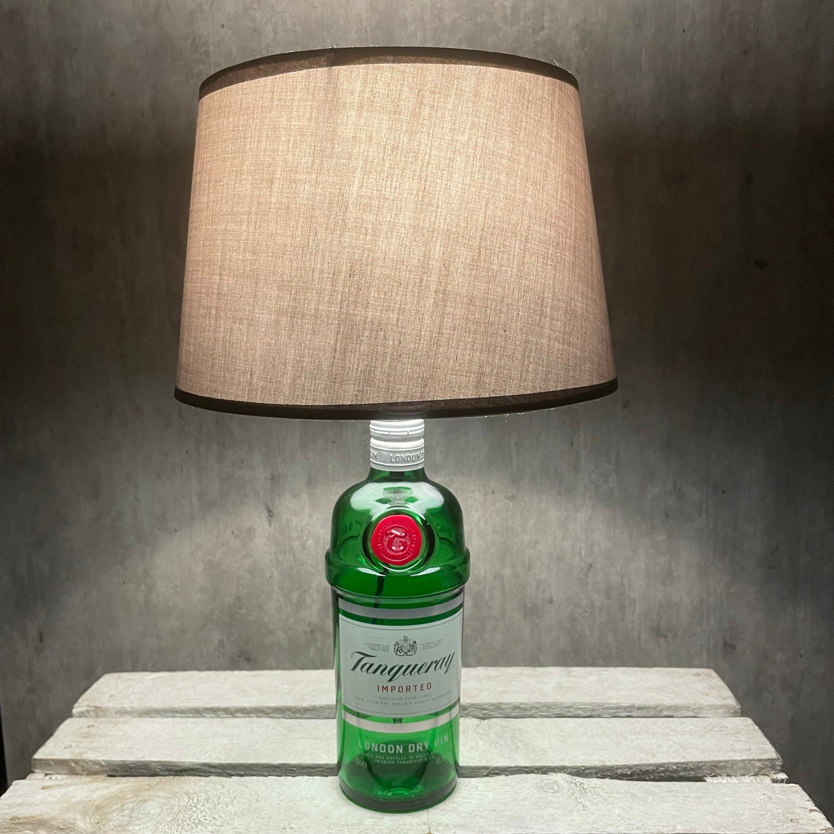 Tanqueray Ginflasche als leuchtende Tischlampe mit hellem Stoffschirm auf Holztisch