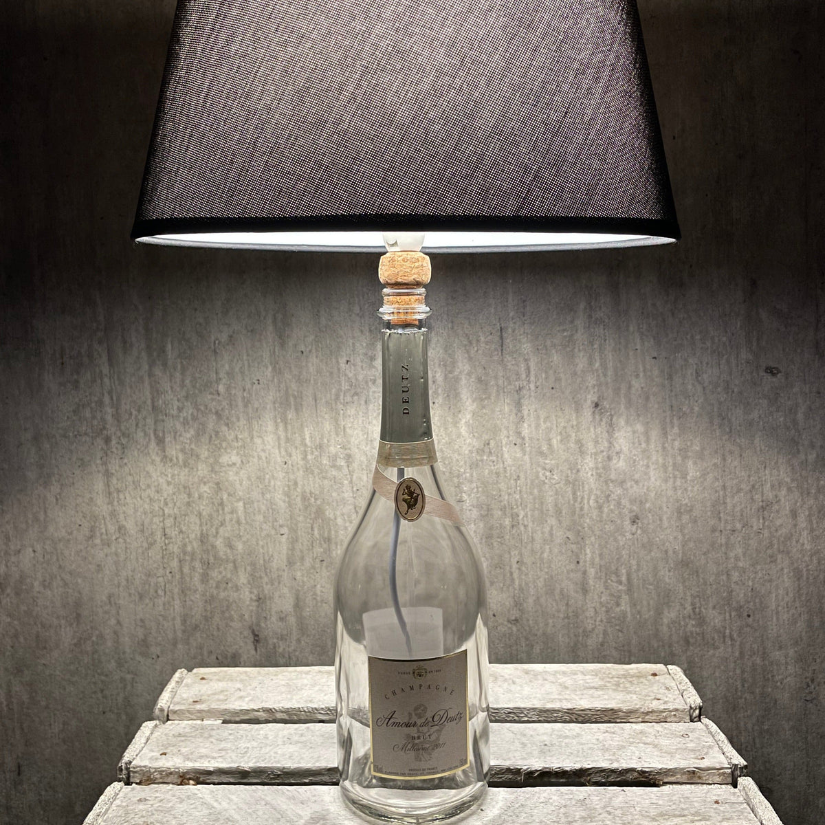Amour de Deutz Champagnerflasche als Tischlampe mit dunklem Stoffschirm vor grauem Hintergrund